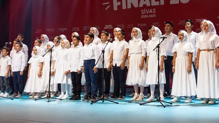 Beyaz kıyafetli çocuk ve gençler, mikrofonların önünde sahnede sıra halinde duruyor; arkada Sivas yazısı görülüyor.