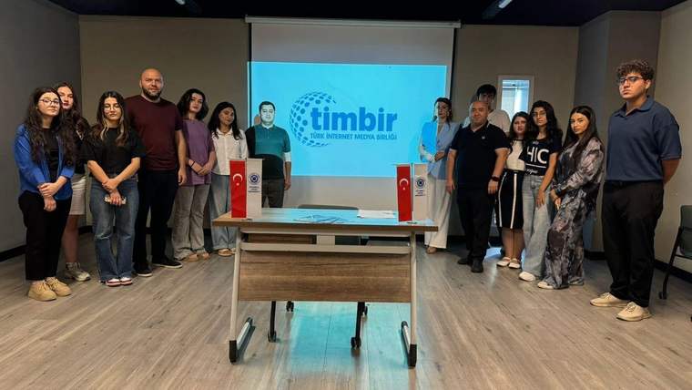 Toplantı salonunda TİMBİR logosunun yansıtıldığı ekran önünde duran kalabalık grup, masanın çevresinde poz veriyor.