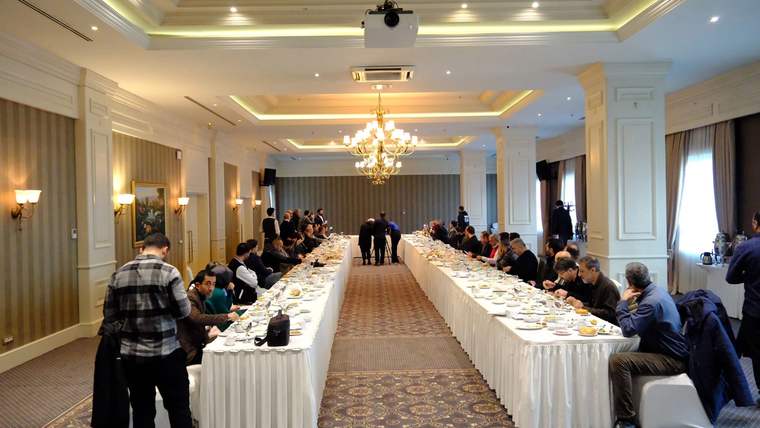 Avizeli bir salonda, uzun masalarda oturan ve yemek yiyen kalabalık bir grup görülüyor.
