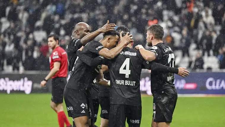 Yağmur altında siyah formalı futbolcular sahada birbirine sarılırken, kırmızı formalı oyuncu arkada duruyor.