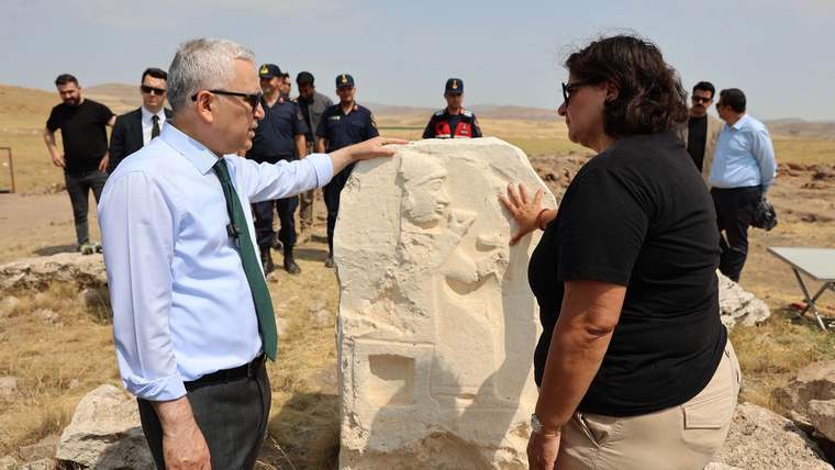 Açık arazide, kabartmalı taş eserin yanında duran iki kişi ve arkalarında bekleyen üniformalı grup görülüyor.