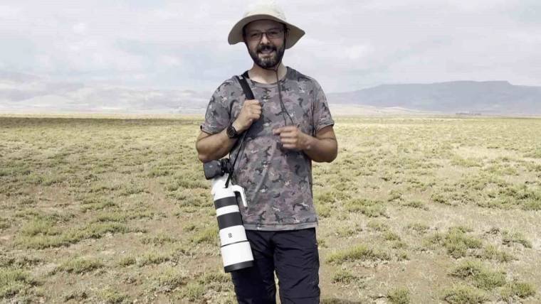 Şapkalı ve gözlüklü bir adam, kayalık tepelerin görüldüğü açık arazide fotoğraf makinesiyle duruyor.