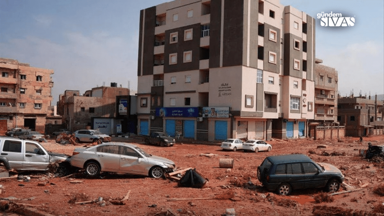 Kırmızı toprakla kaplı alanda hasarlı araçlar, molozlar ve önünde dükkânlar bulunan apartmanlar görülüyor.