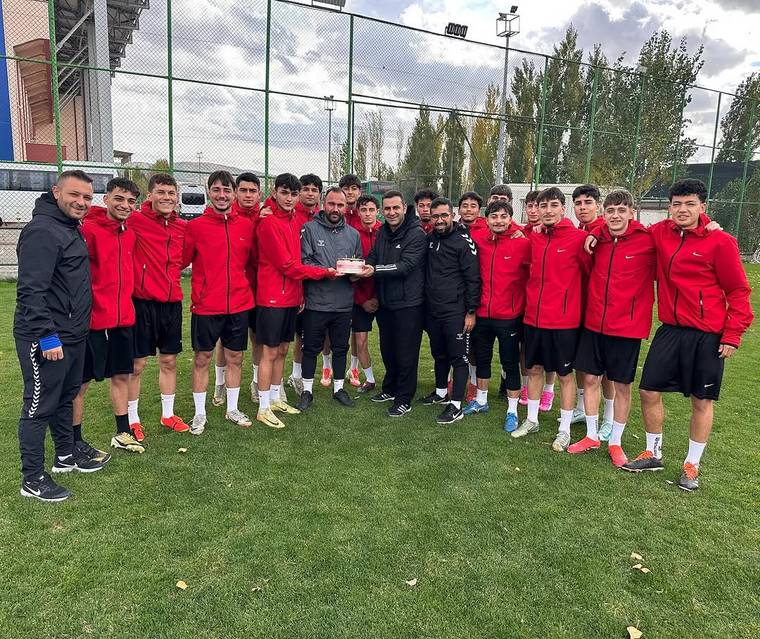 Çim sahada kırmızı eşofmanlı bir grup genç, ortalarında pasta tutan kişilerle birlikte poz veriyor.