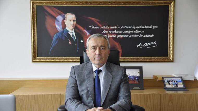 Masa başında oturan takım elbiseli bir kişi, arkasındaki duvarda çerçeveli portre ve yazılı tabloyla görülüyor.