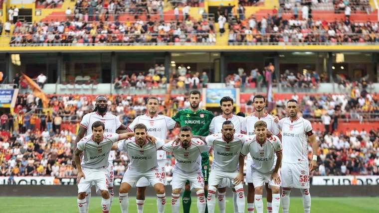 Yeşil çim sahada beyaz formalı futbolcular takım fotoğrafı için dizilirken tribünlerde seyirciler görülüyor.