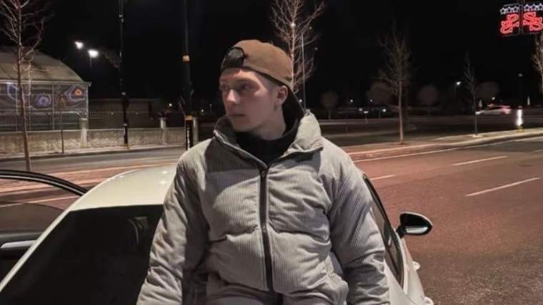 Geceleyin beyaz Volkswagen otomobilin arkasında oturan, gri eşofmanlı ve şapkalı bir kişi görülüyor.