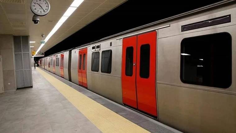 Metro istasyonunun peronunda, kırmızı kapılı gümüş renkli bir tren duruyor; duvarda analog saat görülüyor.