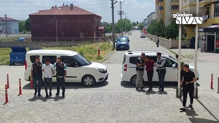 İki araç, bir grup insanın etrafında duruyor; arka planda binalar ve ağaçlar görünmekte.
