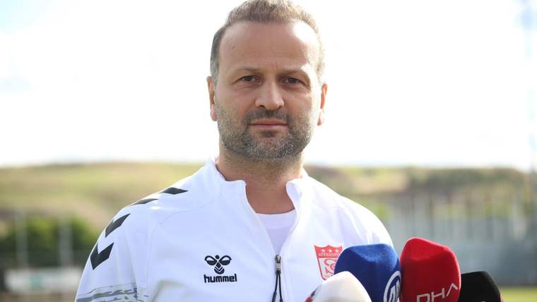 Beyaz spor kıyafetli bir kişi, önündeki DHA ve İHA logolu mikrofonlara açık havada konuşuyor.