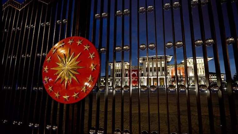 Siyah demir parmaklıklar önünde kırmızı dairesel arma ve ışıklandırılmış bir bina görülüyor.