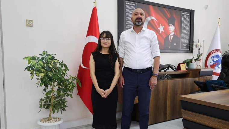 Türk bayrağının önünde, bir kadın ve bir erkek yan yana duruyor. Kadın siyah elbise giymekte, erkek ise beyaz gömlek ve lacivert pantolon giymekte.