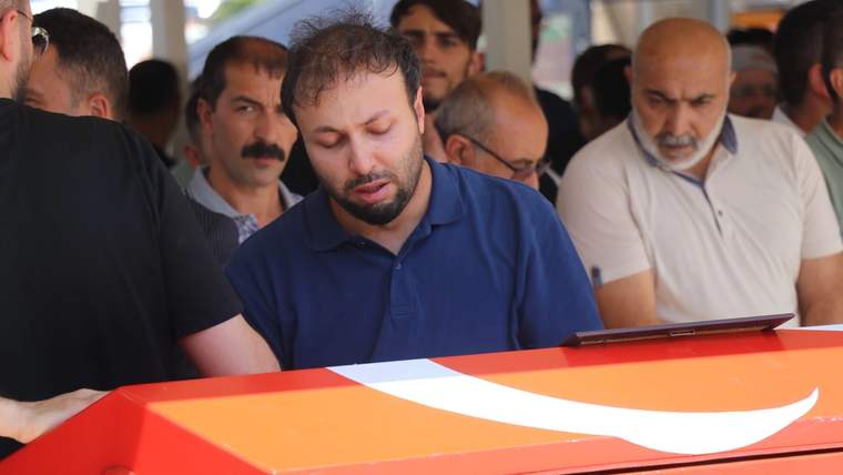 Turuncu-kırmızı renkli, üzerinde beyaz ay ve yıldız bulunan tabutun çevresinde insanlar duruyor.