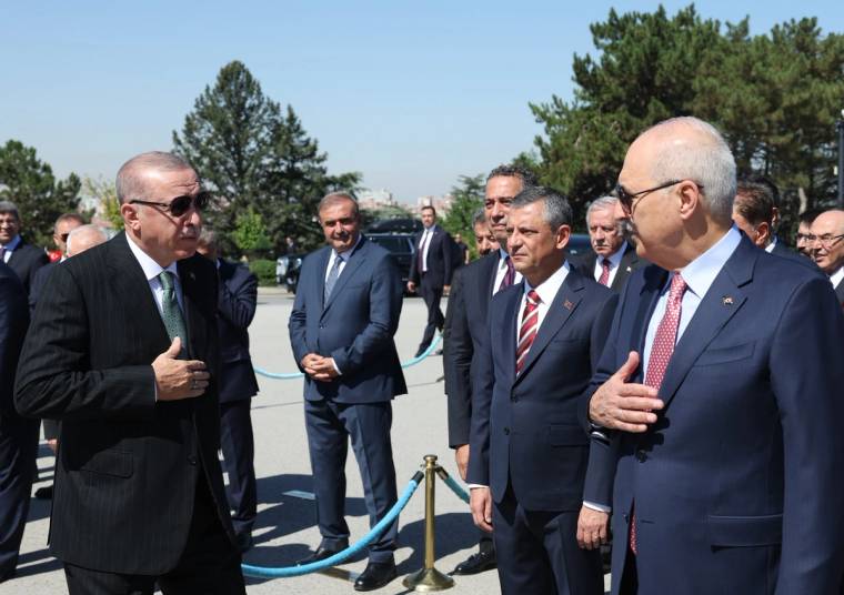 Açık havada takım elbiseli bir grup erkek, mavi halatlı bariyerin yanında ayakta duruyor.