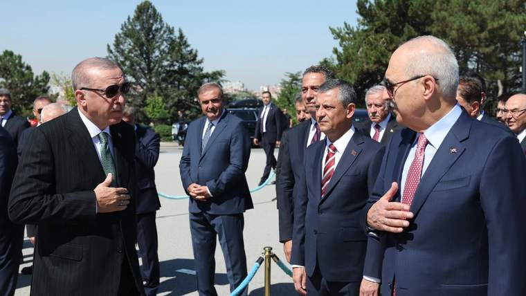 Açık havada takım elbiseli bir grup erkek, mavi halatlı bariyerin yanında ayakta duruyor.