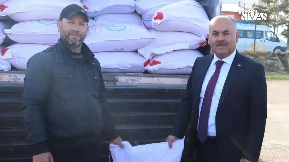Bir kişi, arka planda torbalarda arpa ile birlikte başka bir kişiyle poz veriyor.