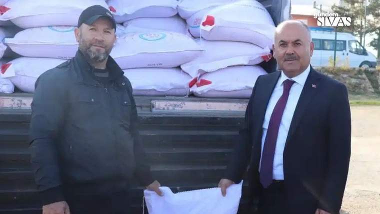 Bir kişi, arka planda torbalarda arpa ile birlikte başka bir kişiyle poz veriyor.