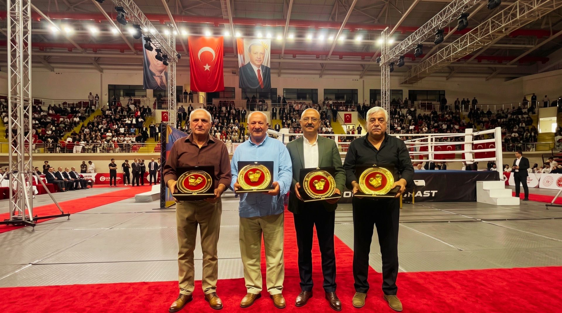 Spor salonunda dört kişi, ellerinde kırmızı kuşaklı altın renkli ödül plakalarıyla yan yana duruyor.