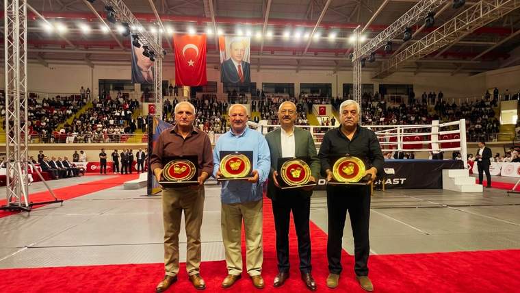 Spor salonunda dört kişi, ellerinde kırmızı kuşaklı altın renkli ödül plakalarıyla yan yana duruyor.