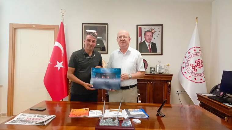 Bir kişi, masada oturan başka bir kişiye hediye verirken gülümsüyor; arka planda Türk bayrağı ve portreler var.