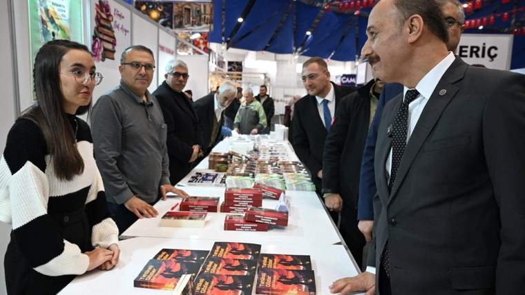 Bir stantta kitaplar sergileniyor, bir kişi kitapları inceliyor, yanındaki kişilerle sohbet ediliyor.