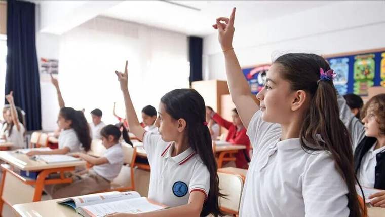 Sınıfta oturan öğrencilerden bazıları ellerini kaldırırken, ön plandaki iki öğrenci ders kitaplarının yanında görülüyor.