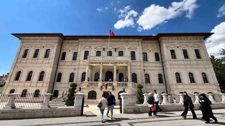 Taş cepheli tarihi müze binasının önünde Türk bayrağı ve yürüyen ziyaretçiler görülüyor.