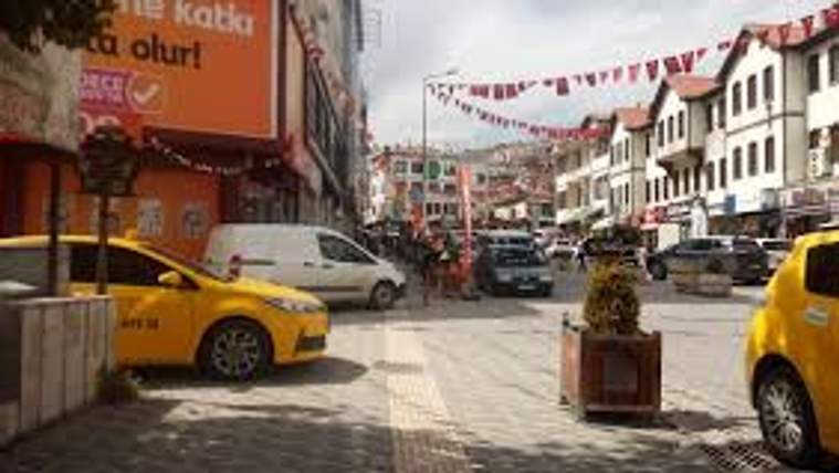 Cadde üzerinde sarı ve beyaz araçlar, sıra binalar, saksıdaki ağaç ve asılı bayrak süslemeleri görülüyor.