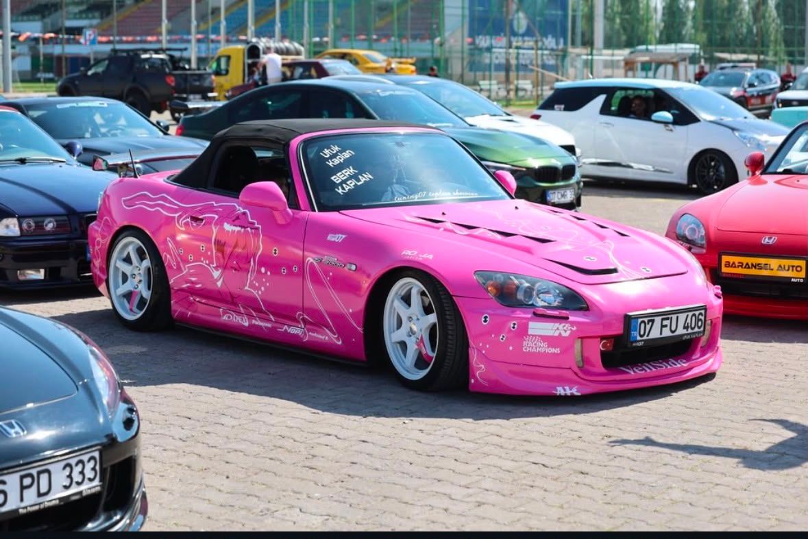 Otoparkta, aralarında pembe modifiye spor otomobilin de bulunduğu çeşitli araçlar yan yana duruyor.