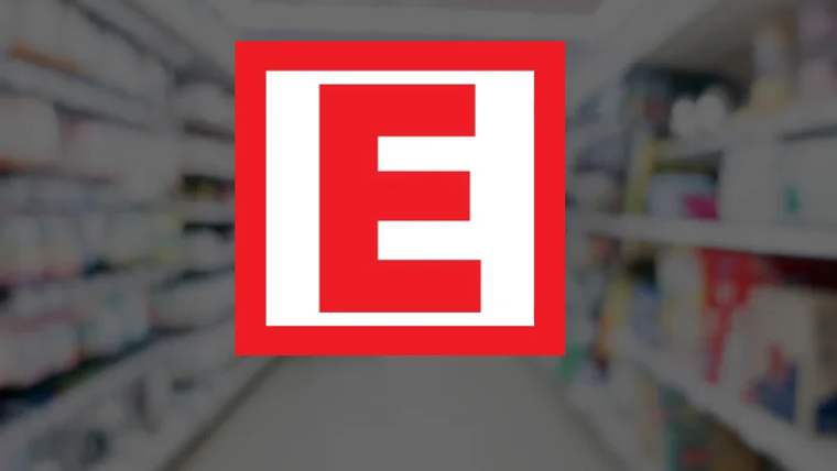 Bulanık eczane raflarının önünde, beyaz çerçeveli kırmızı kare içinde eczane sembolü görülüyor.