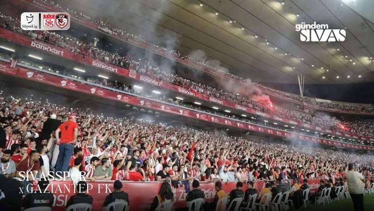 Stadyum tribünlerinde kırmızı beyaz bayraklar ve atkılar taşıyan kalabalık taraftar grubu görülüyor.