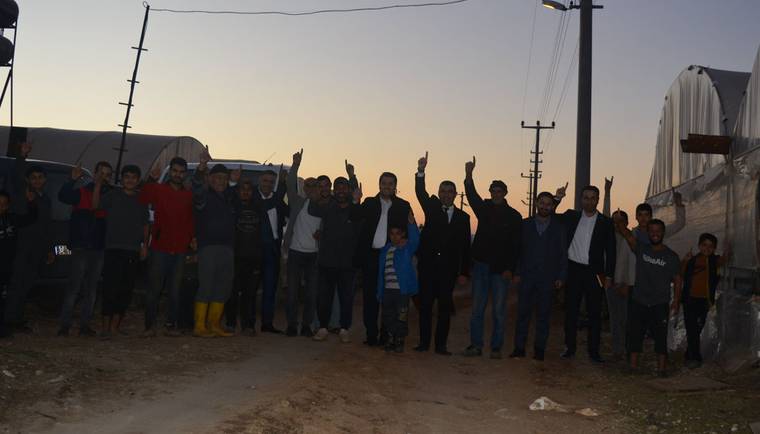 Toprak yolda bir arada duran kalabalık, bazı kişiler ellerini havaya kaldırırken arkada örtülü yapılar görülüyor.