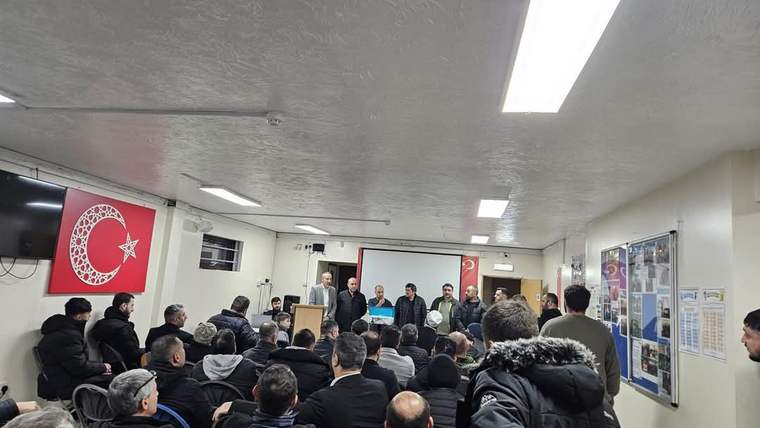 Bir grup insan, bir salonda sahneye bakan şekilde oturmuş. Sahne önünde birkaç kişi duruyor.
