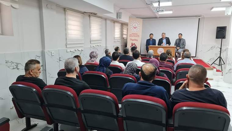 Toplantı salonunda oturan katılımcılar, önde konuşan üç kişinin bulunduğu kürsüye bakıyor.