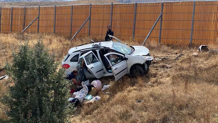 Kuru otların arasında yoldan çıkmış beyaz otomobilin çevresinde birkaç kişi ve açılmış kapılar görülüyor.