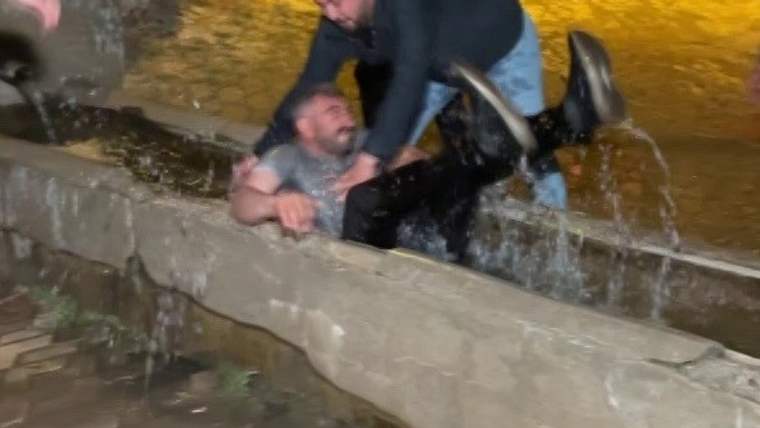 Gece çekiminde, beton su kanalında oturan bir adamın yanında eğilen başka bir adam ve akan su görülüyor.