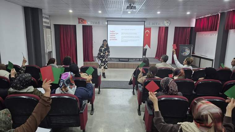 Seminer salonunda kürsü önünde konuşan bir kişi ve ellerinde renkli kartlar tutan katılımcılar görülüyor.