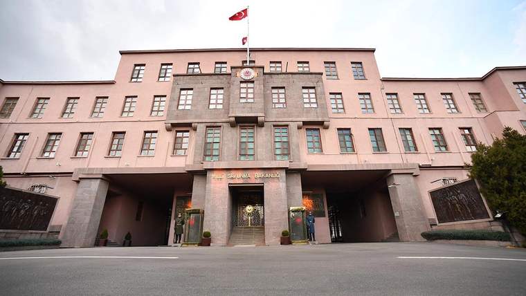 Türk bayrağı bulunan pembe renkli çok katlı binanın sütunlu ana girişi ve önündeki geniş asfalt alan görülüyor.
