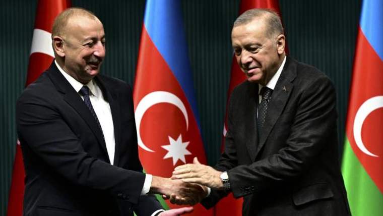 Azerbaycan ve Türkiye bayrakları önünde iki takım elbiseli kişi el sıkışıyor.