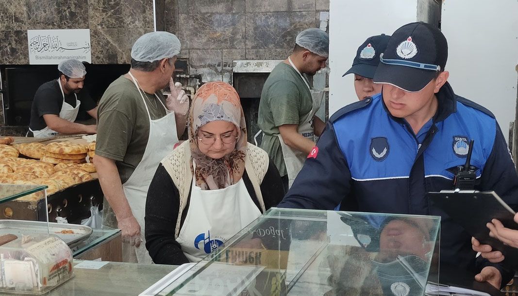 Üniformalı iki görevli, bone ve önlük takan çalışanlarla birlikte tezgâhın başında denetim yapıyor.