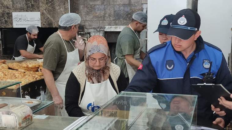 Üniformalı iki görevli, bone ve önlük takan çalışanlarla birlikte tezgâhın başında denetim yapıyor.