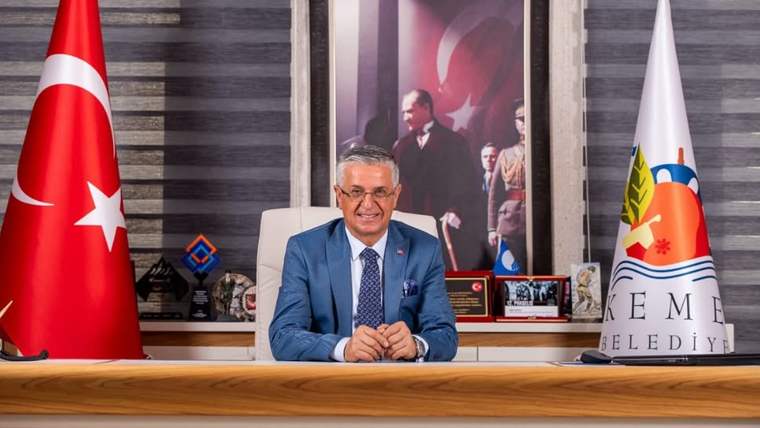 Masa başında oturan bir kişi, Türk bayrağı ve belediye logolu bayrakların arasında kameraya bakıyor.