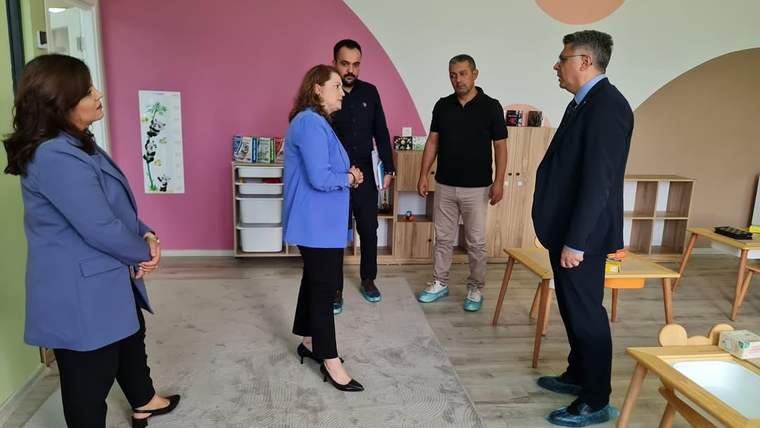 Bir odada, dört kişi arasında konuşma yapılıyor; arka planda oyuncaklar ve çocuk masaları var.