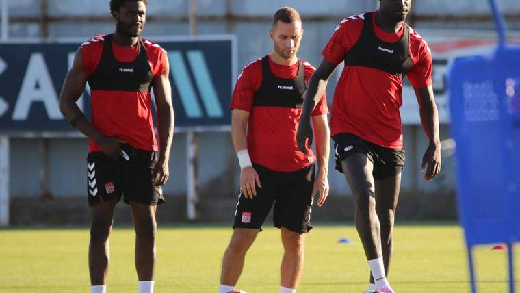 Antrenman sahasında kırmızı formalar giyen üç futbolcu, toplarla birlikte yürürken görülüyor.