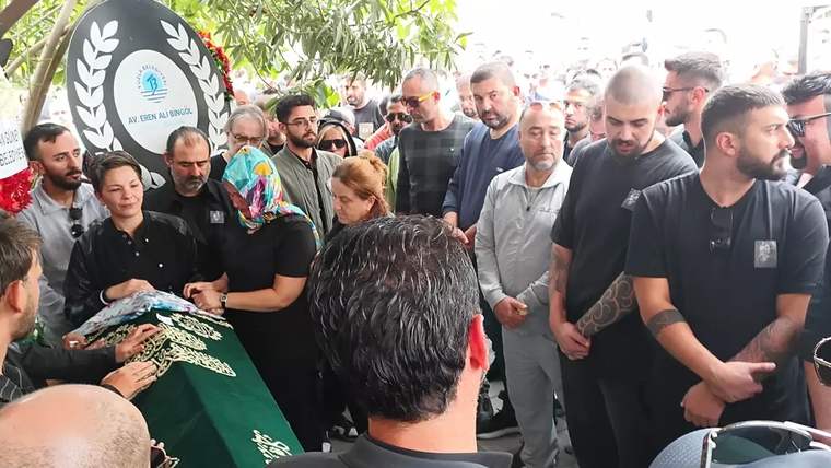 Yeşil örtüyle kaplı cenaze tabutu çevresinde, açık alanda toplanan kalabalık ve bir çelenk görülüyor.