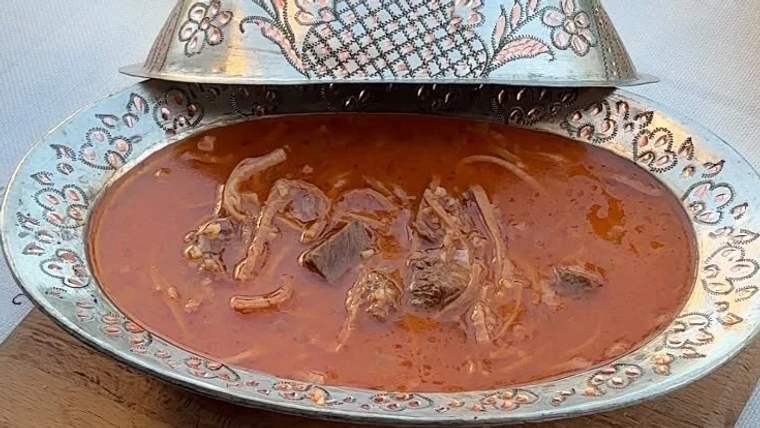 Desenli metal bir kâsede, kırmızı renkli et parçaları ve soğan bulunan sulu yemek sunuluyor.