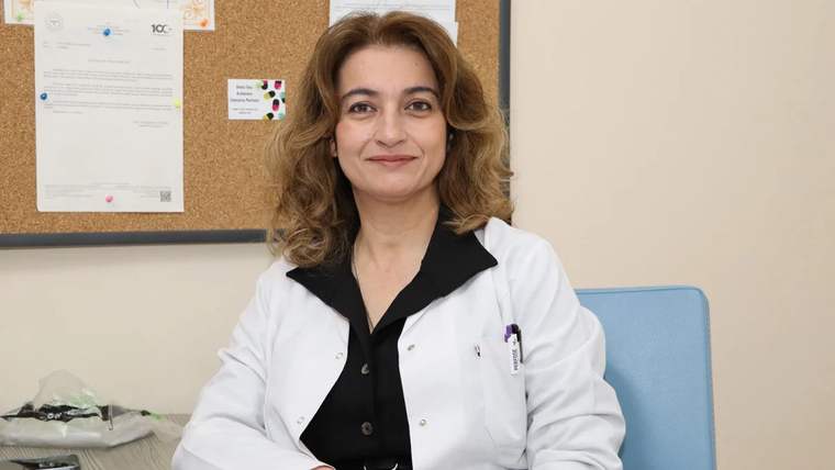 Beyaz önlük giymiş bir kadın, arkasında duyuru panosu bulunan bir odada oturuyor.