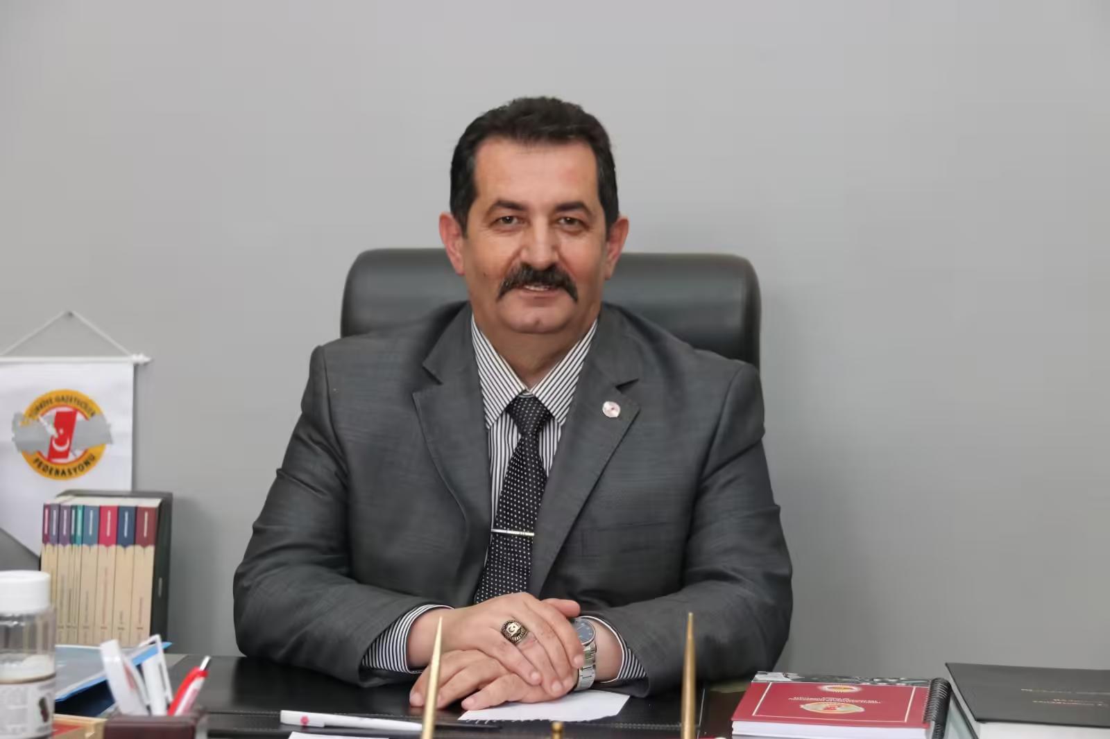 Bir masa başında oturan, gri takım elbise giymiş bir kişi, arkasında beyaz duvar ve masa üzerinde dosyalar var.