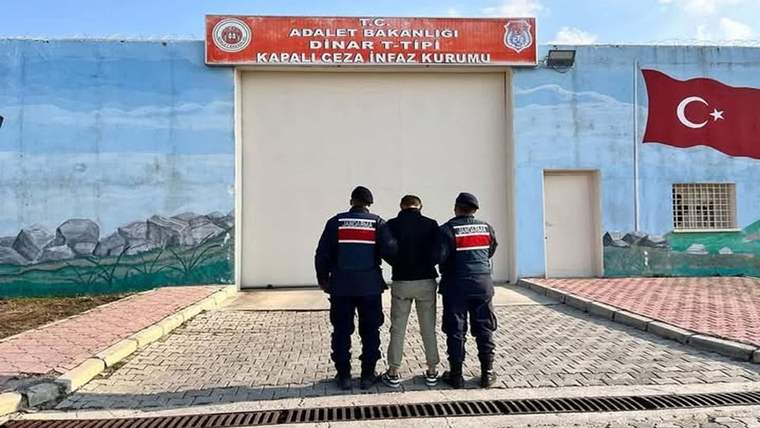 Üzerinde Dinar T Tipi Kapalı Ceza İnfaz Kurumu yazan girişte iki üniformalı görevli bir kişiye eşlik ediyor.