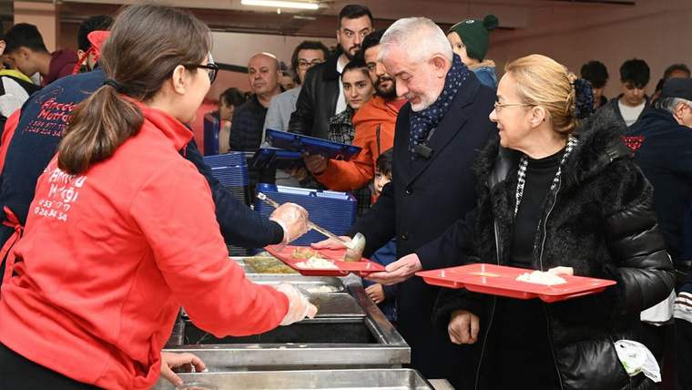 Yemek dağıtım noktasında kırmızı tepsiler taşıyan kalabalık, servis yapan görevlilerden yemek alıyor.
